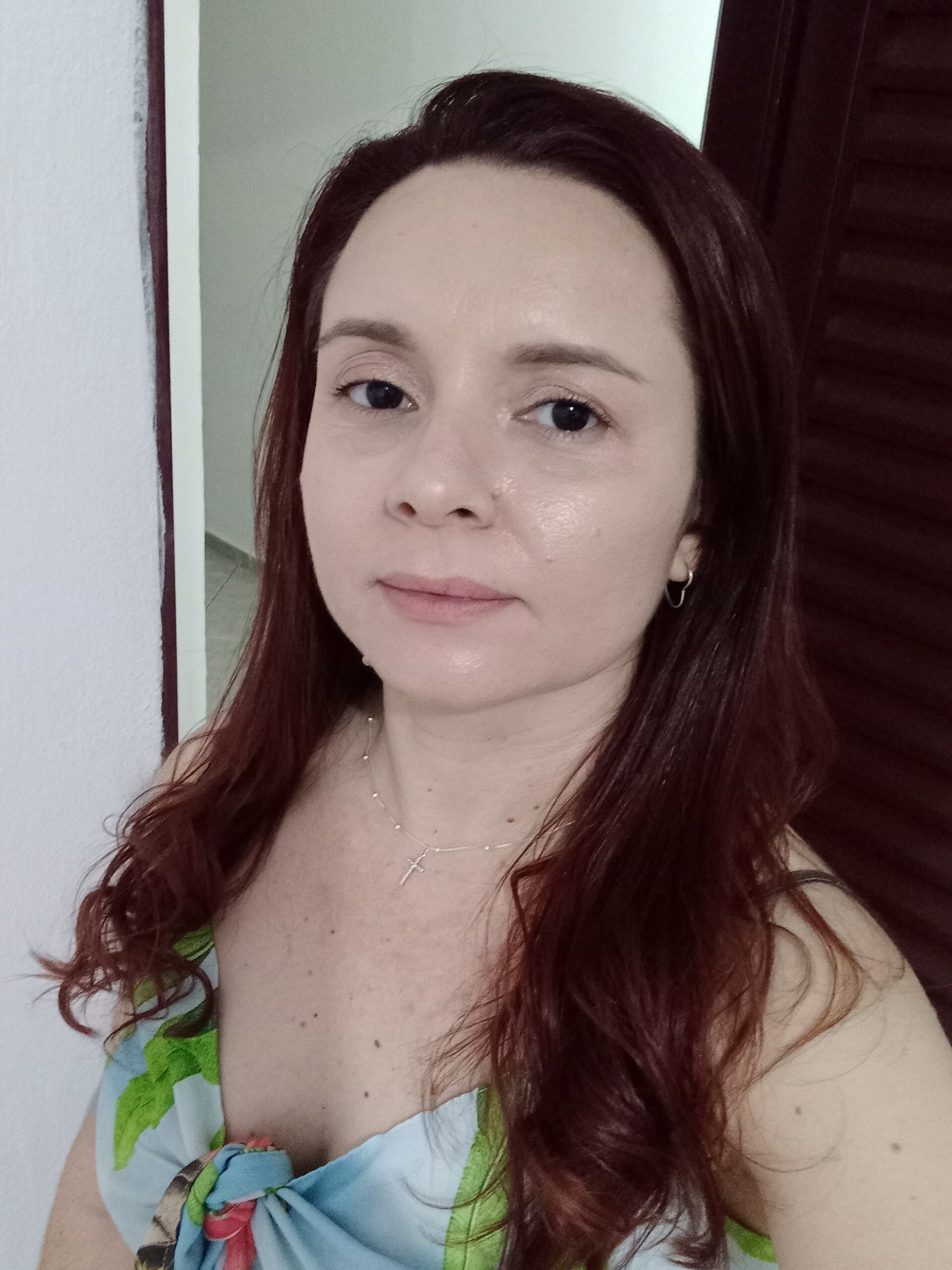 Uma mulher de cabelos longos, castanho-avermelhados, olha diretamente para a câmera. Ela usa uma blusa de alças com estampa floral em tons de azul e verde, um colar com um pingente de cruz e pequenos brincos de argola. Ela está em um ambiente interno com uma parede branca e uma porta ou painel de madeira escura ao fundo.