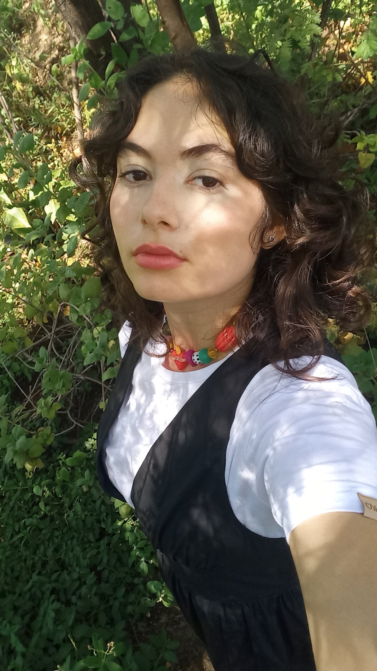Mulher de cabelos escuros e ondulados posa ao ar livre com folhagens verdes ao fundo. Ela veste uma camiseta branca sob um vestido preto de alças e usa um colar colorido com miçangas grandes. A luz do sol cria sombras suaves em seu rosto.