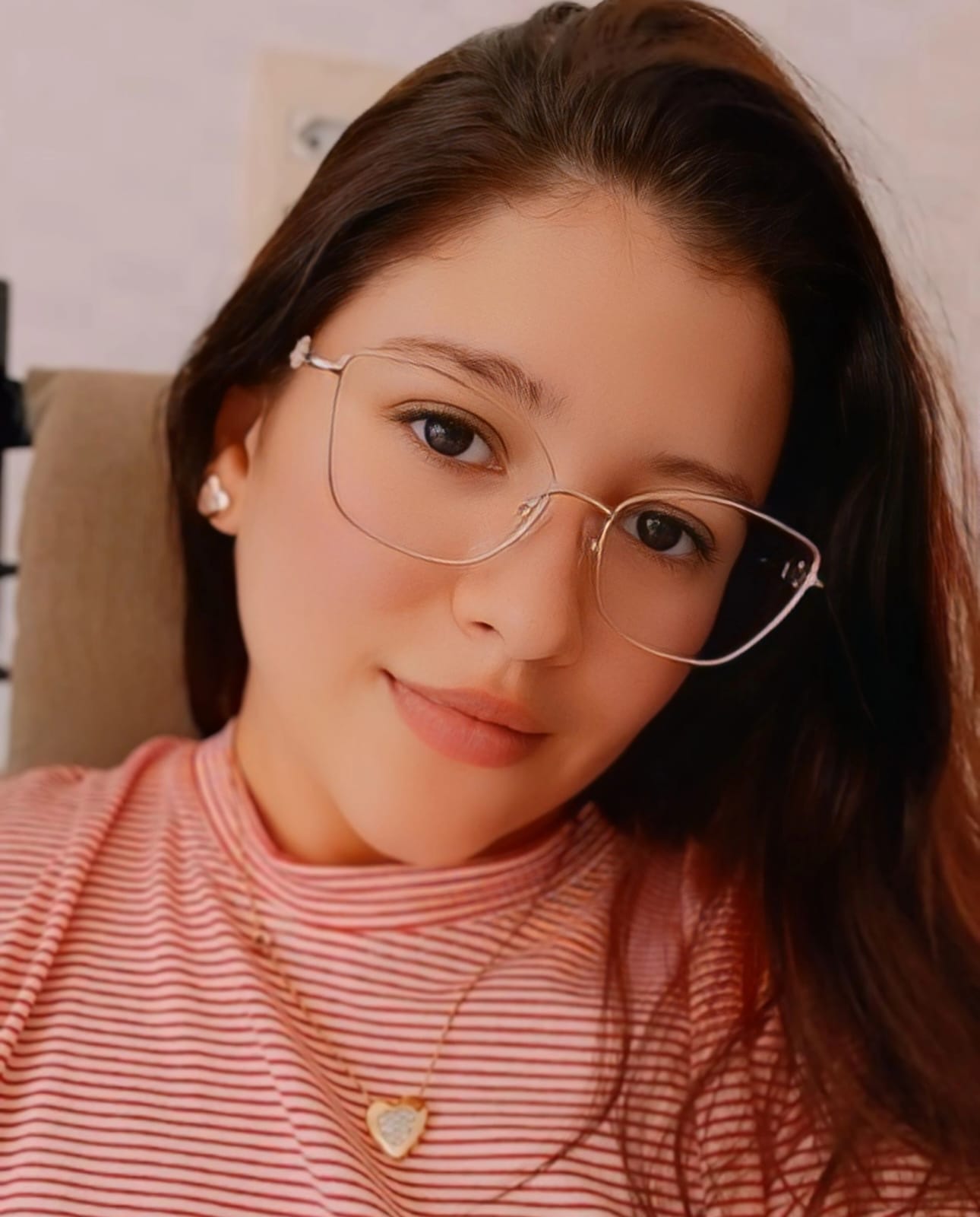 Mulher jovem de pele clara, cabelos longos e castanhos, usando óculos de armação transparente e blusa listrada rosa, olhando para a câmera com expressão serena, em ambiente interno.