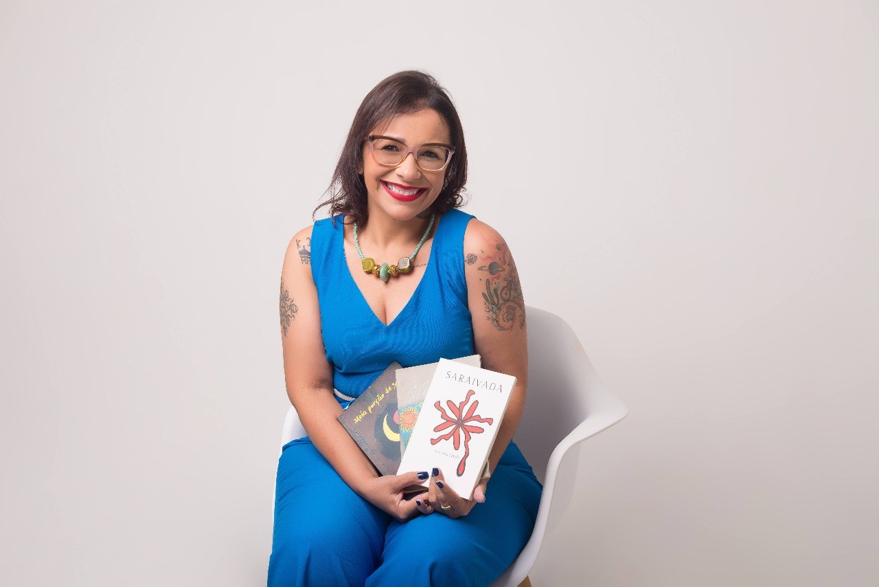 Mulher adulta sentada em uma cadeira branca, sorrindo para a câmera, usando vestido azul, óculos e colar colorido, com tatuagens nos braços, segurando três livros em um estúdio de fundo claro.